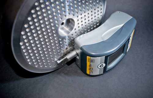 Round metal part with numerous holes and a digital measuring device placed against its surface.