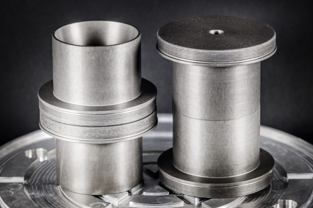 Two precision-turned metal components with flanges, positioned on a metal disc against a dark background.
