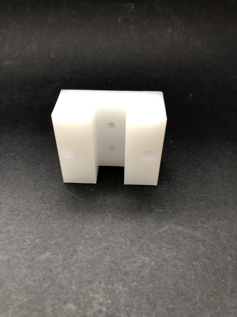 A white U-shaped milled plastic component with visible holes, lying against a dark background.