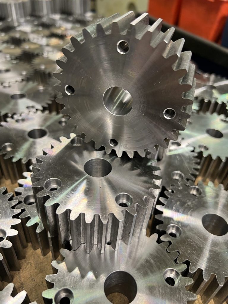 Metal gears with mounting holes and teeth, arranged in a pile against a background of other milled components.