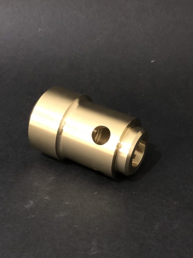 A brass turned cylindrical component with stepped diameters and a cross hole, lying against a dark background.