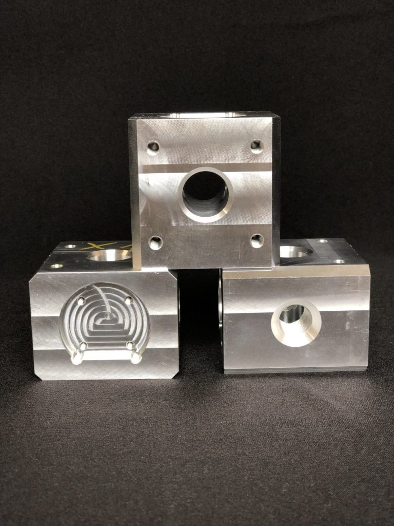 Three milled metal CNC components arranged in a pyramid, with visible holes and grooves against a black background.