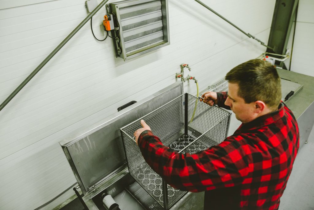 A man in a red-and-black shirt places a steel basket with machined components into a metal washer tank.