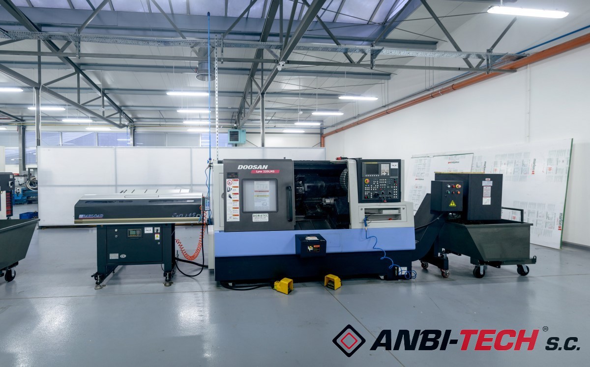 A blue and grey CNC lathe stands in a spacious, bright production hall next to a bar feeder and a chip trolley.