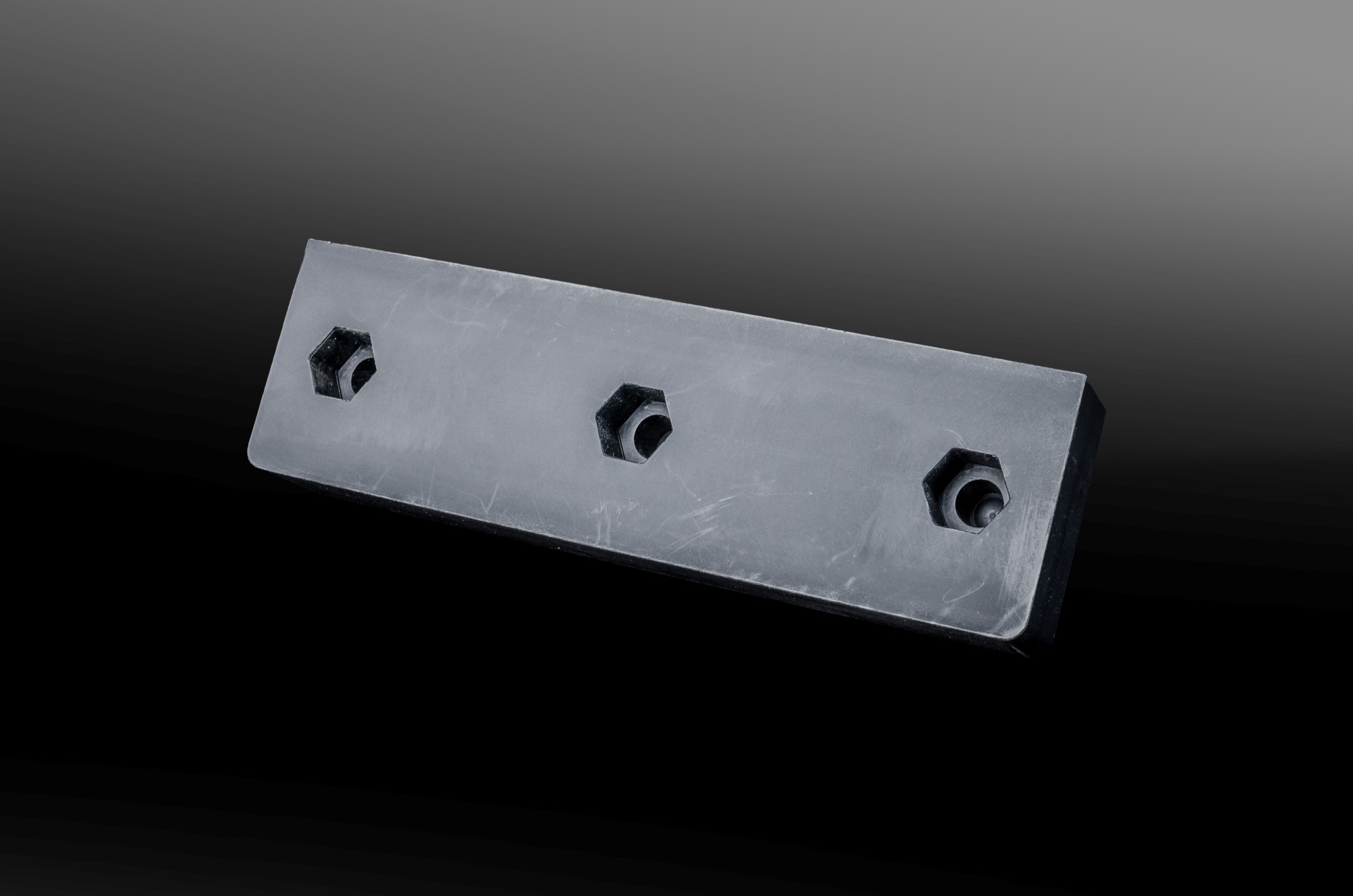 Rectangular black plastic part with three hexagonal recesses at the holes, against a dark background.