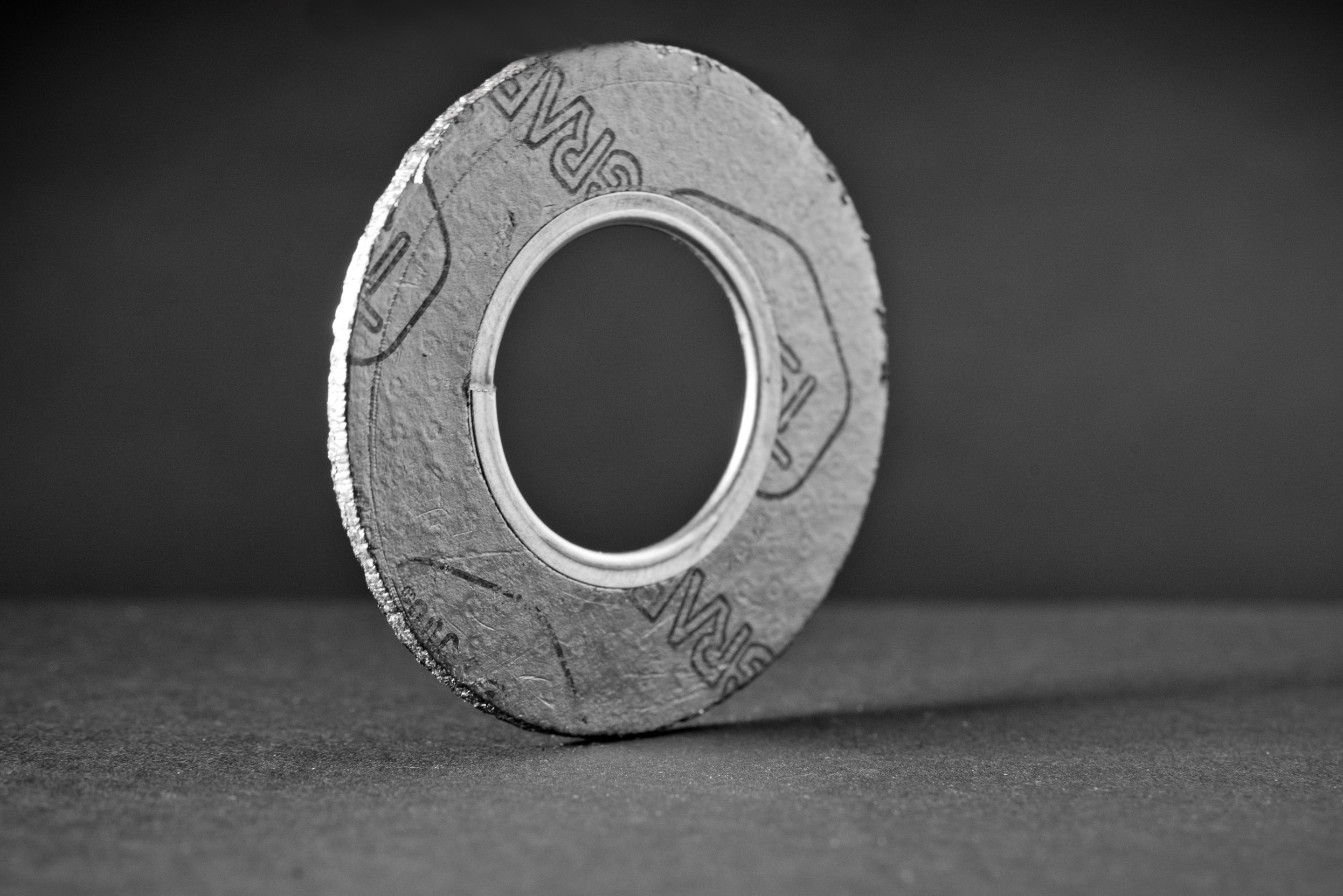 A circular graphite gasket with an inner metal ring, standing upright on a dark surface.