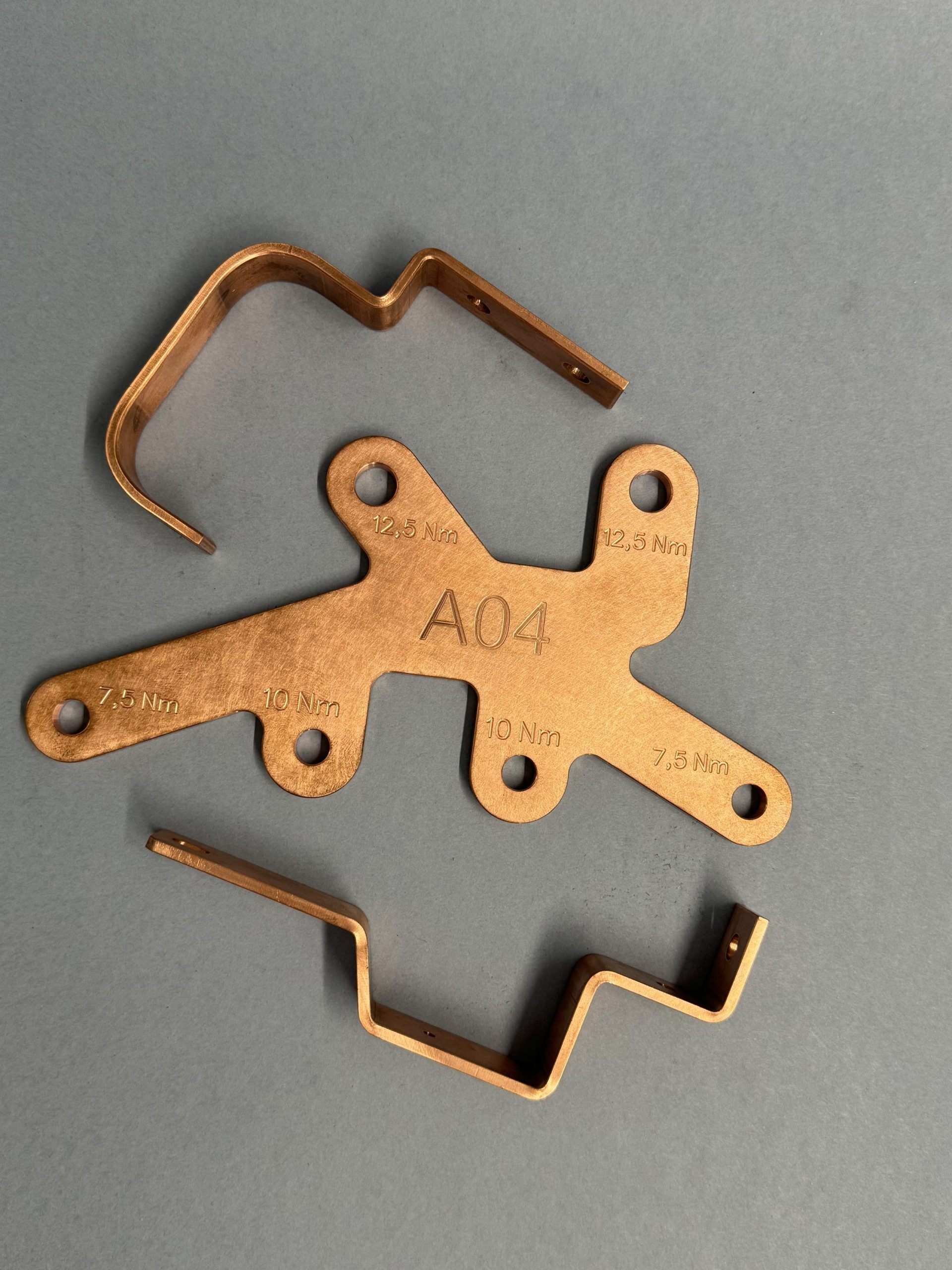 Three copper components on a grey background, including a flat plate marked A04 with Nm markings and two bent profiles.