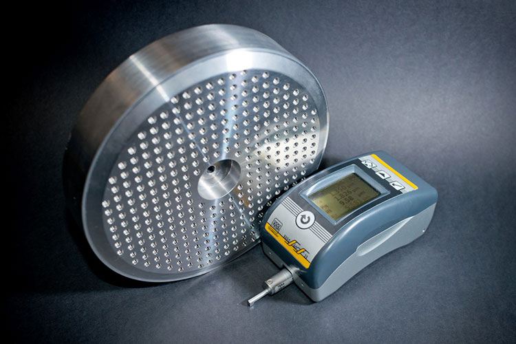 A round metal disc with a dense pattern of holes lying next to an electronic roughness tester on a black background.
