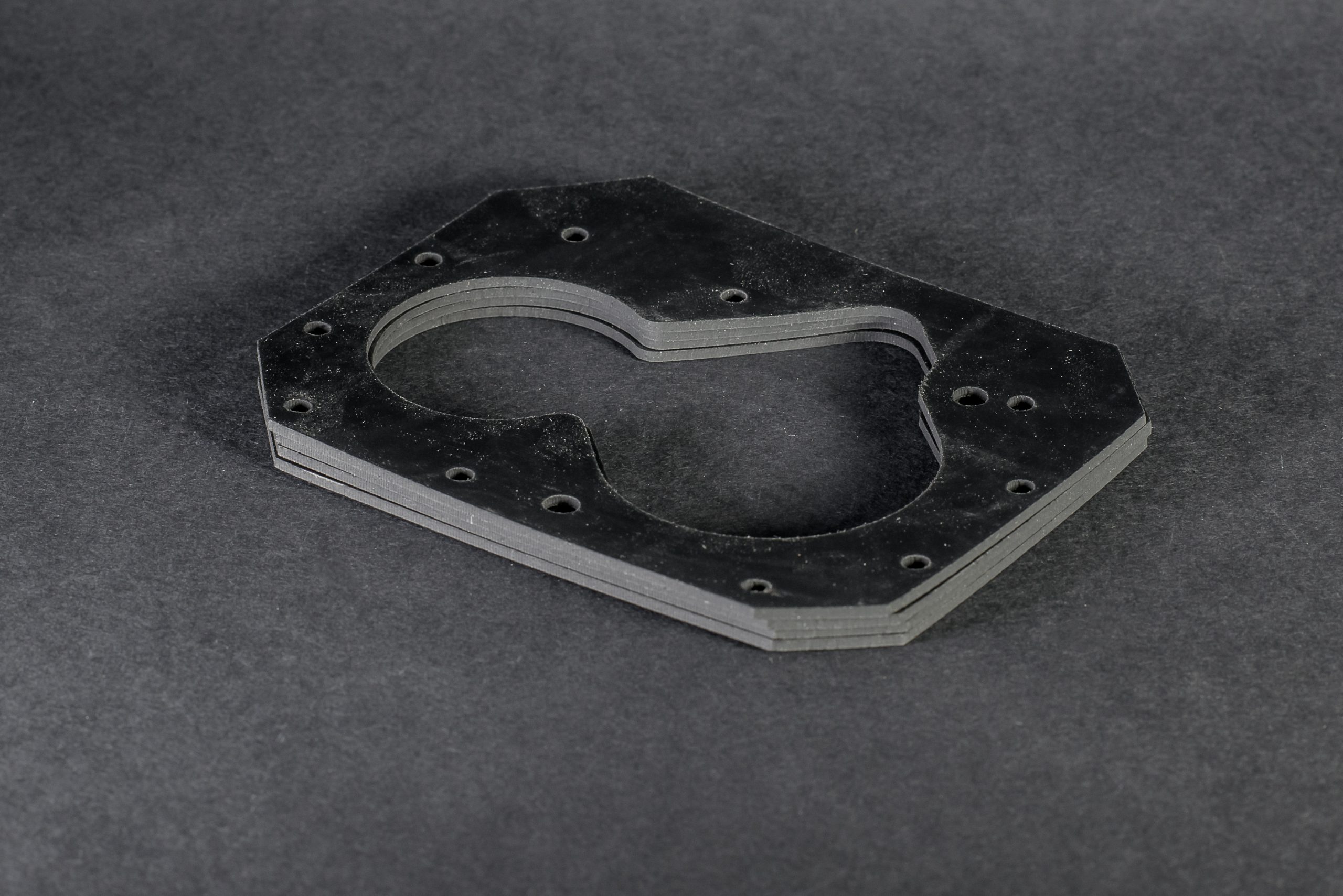 A stack of flat black technical gaskets with irregular outlines and bolt holes on a dark surface.