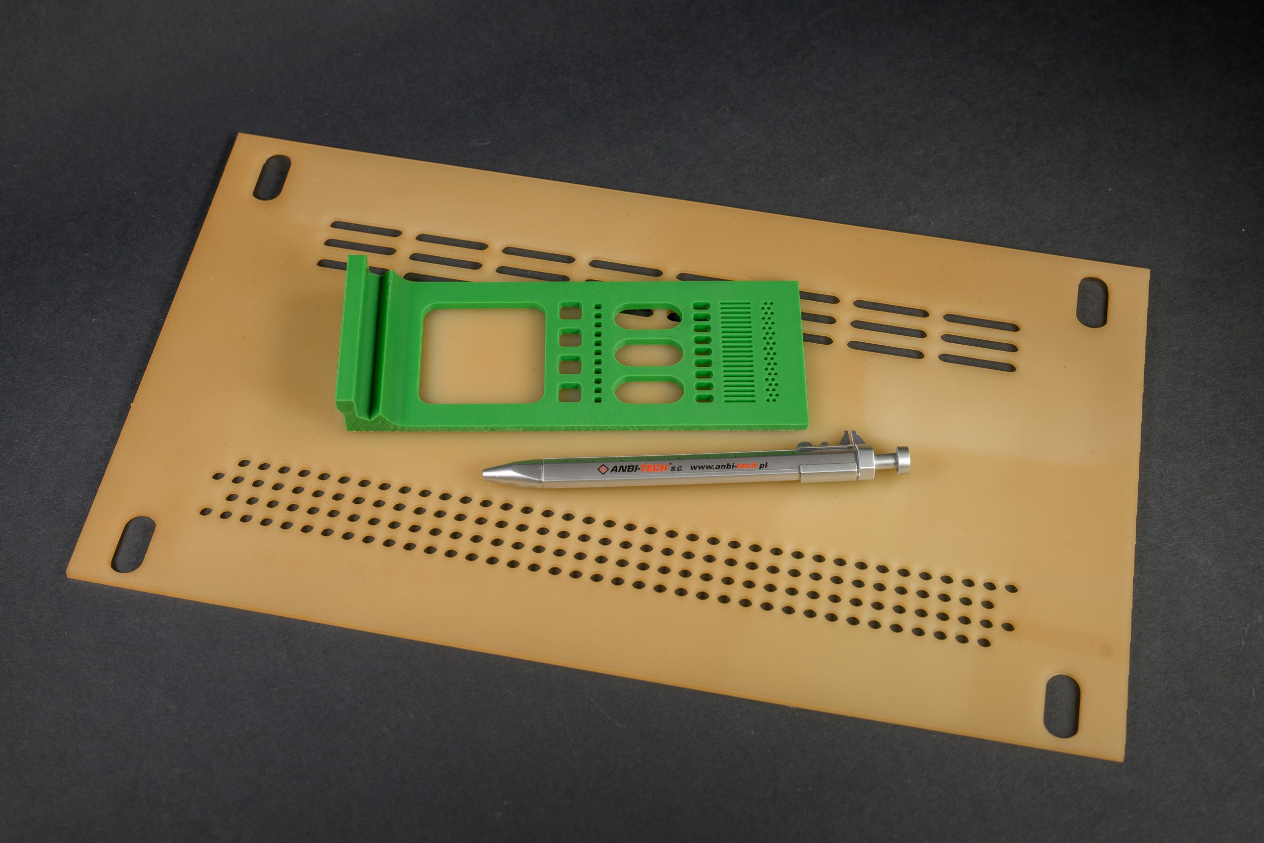 A yellow-brown sheet with elongated and round holes, a green cut-out component and a metal pen on a black background.