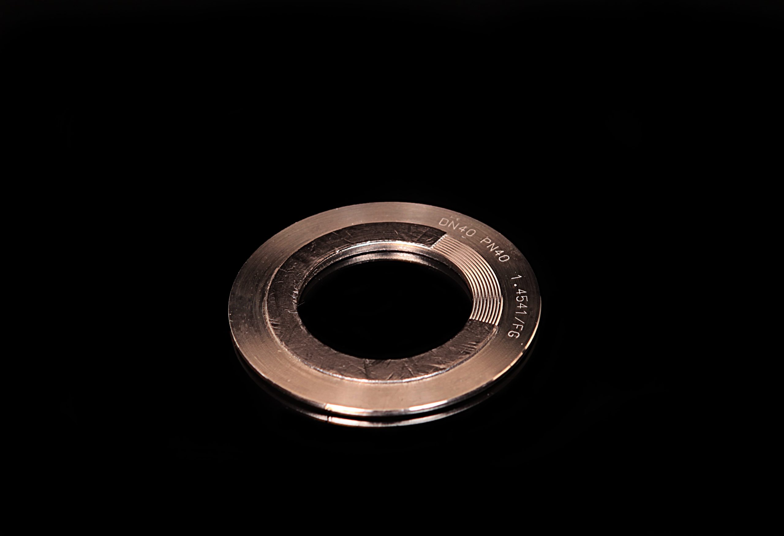 A metal ring gasket with embossed markings, lying on a smooth, black reflective background.