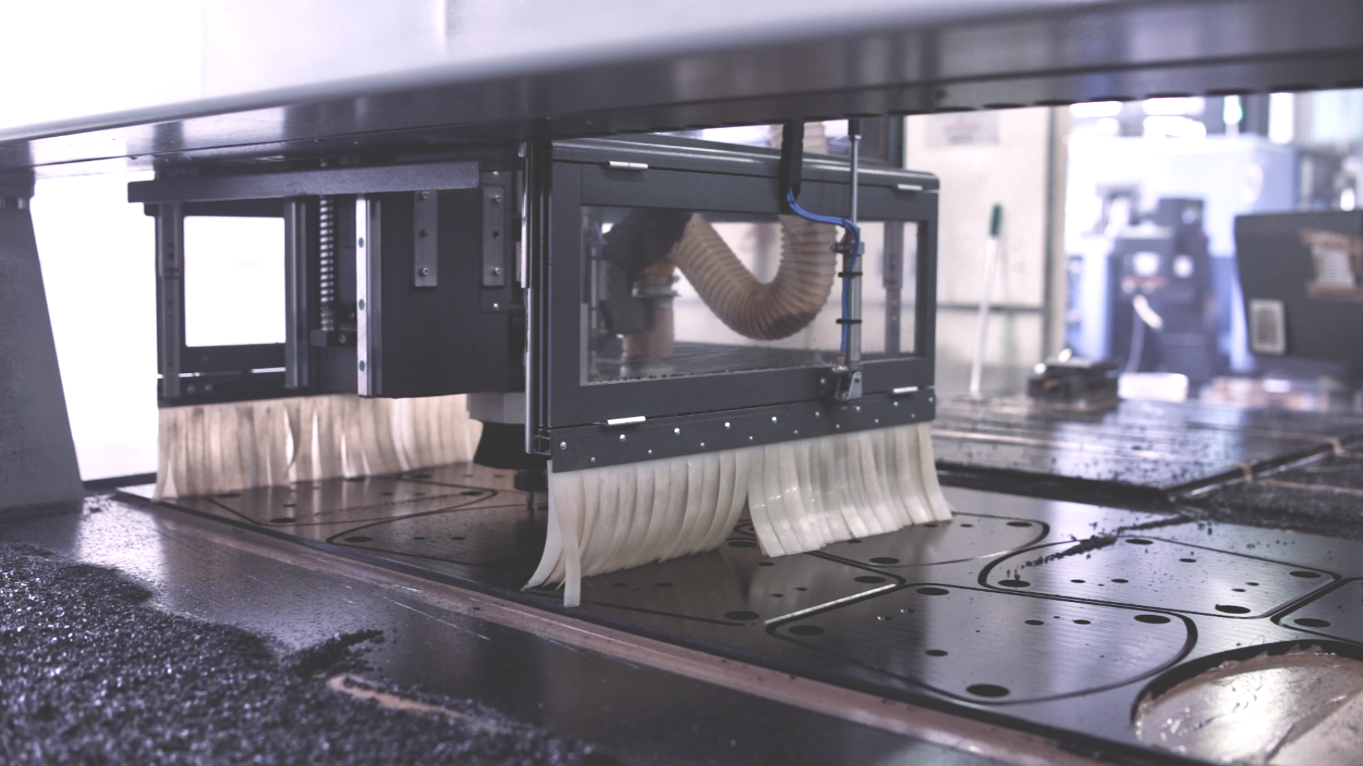 CNC milling head with a protective brush machining a black sheet with cut-out shapes on the work table.