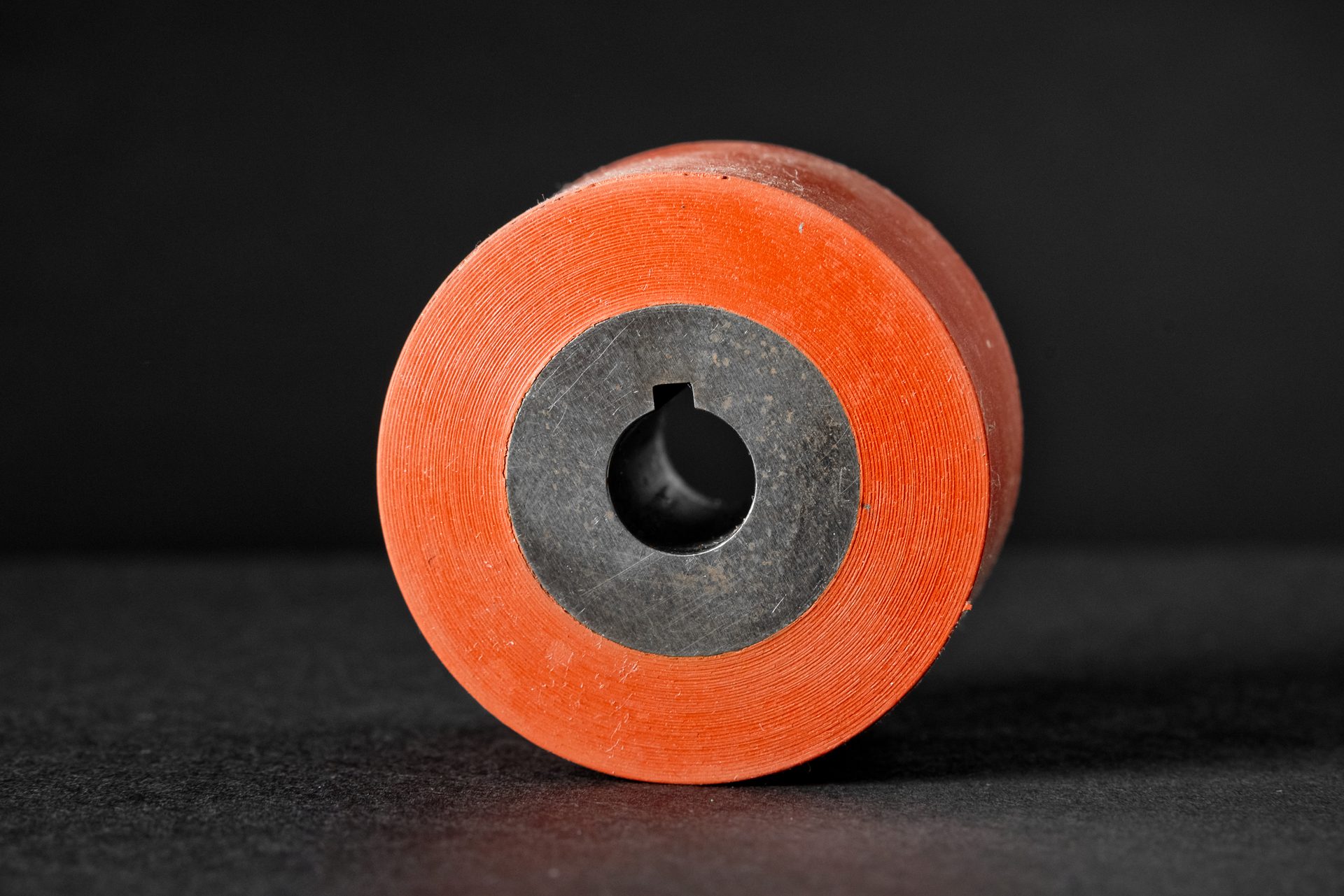 An orange cylinder with a metal core and a central bore for a shaft stands on a smooth black surface.
