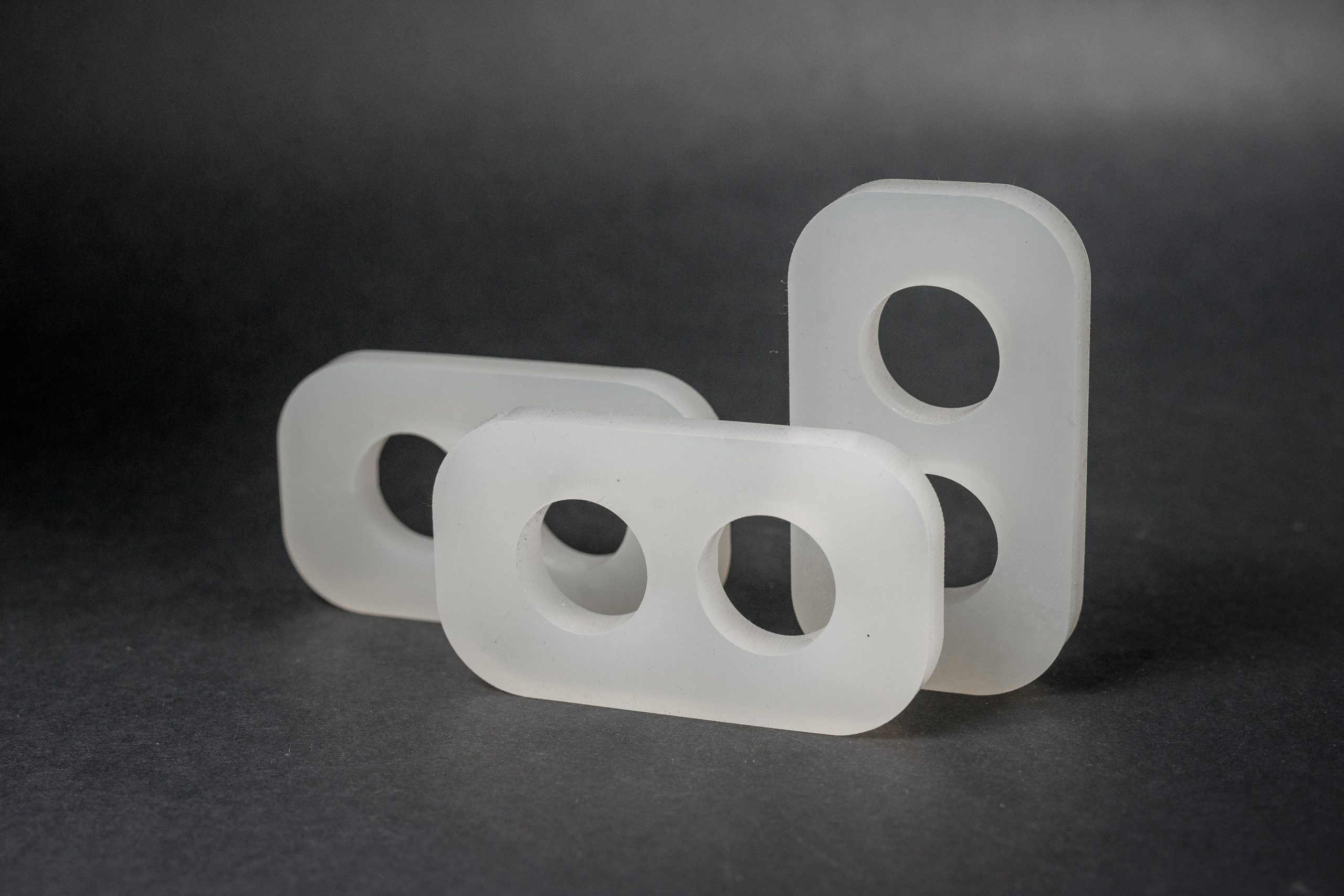 Three rectangular white plastic parts with holes arranged on a dark, matte background.