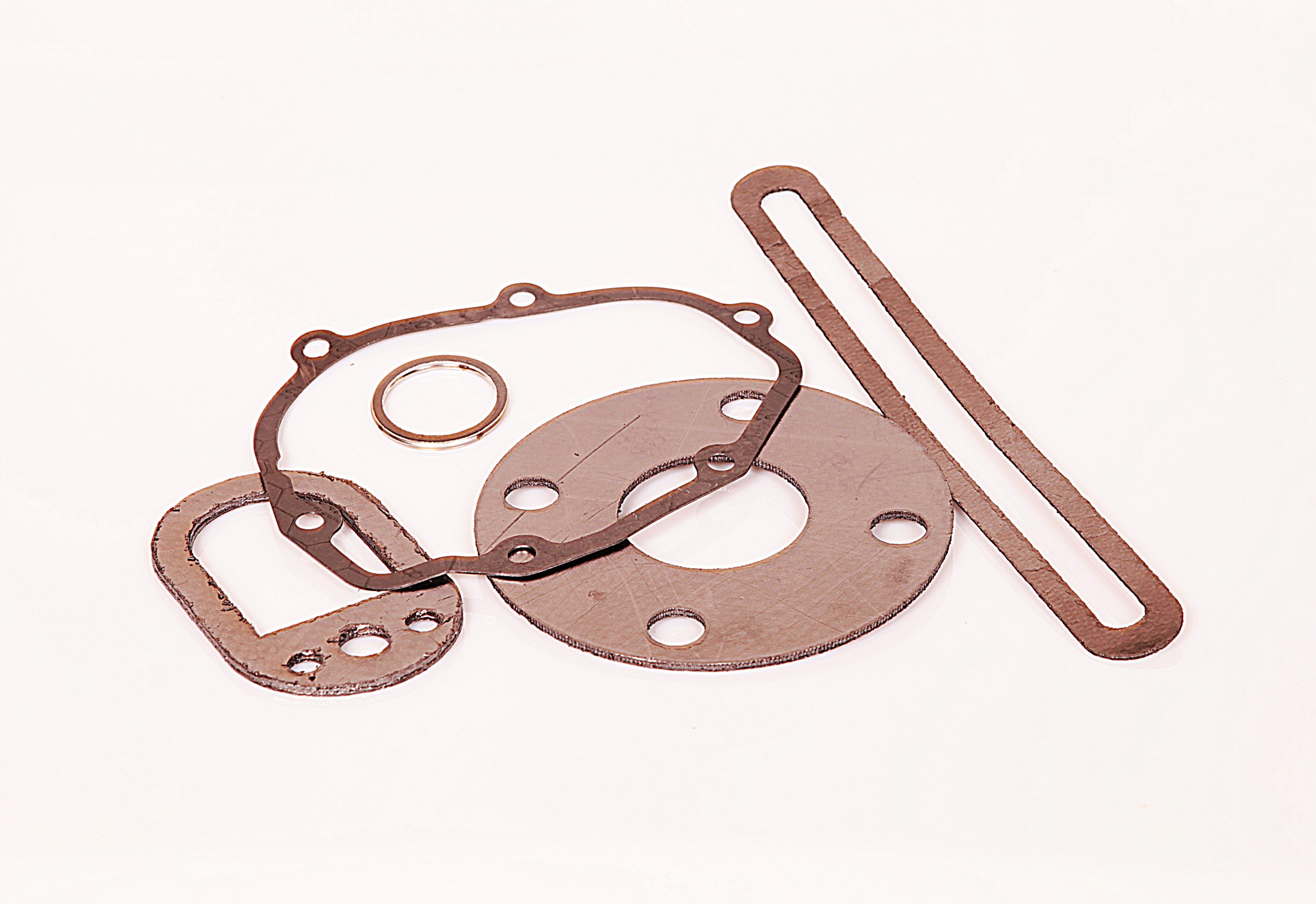 Flat gaskets of various shapes and grey shades arranged on a white background next to a small metal ring.