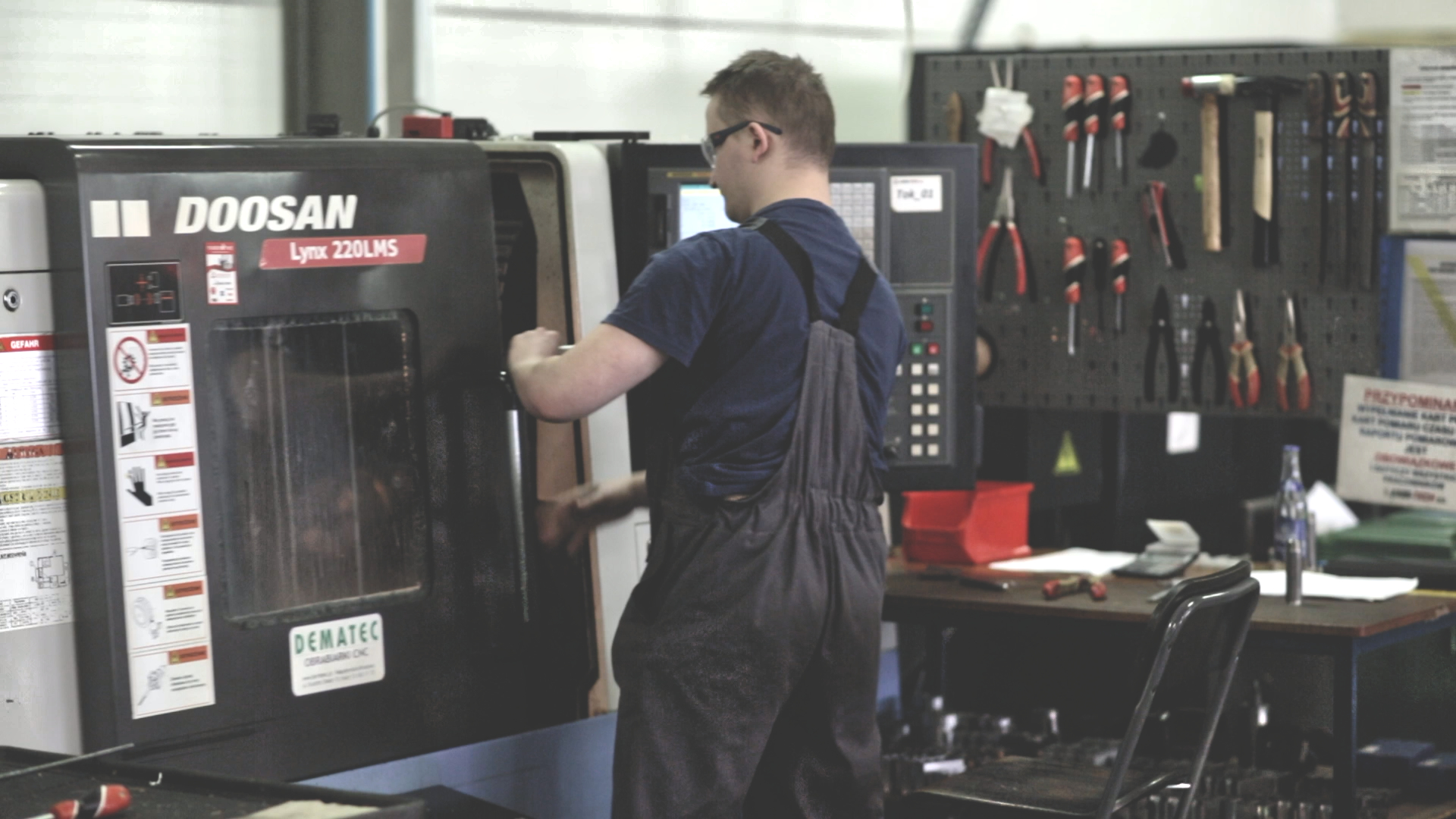 Man in dark dungarees standing by a large black machine, with a tool board visible in the background.
