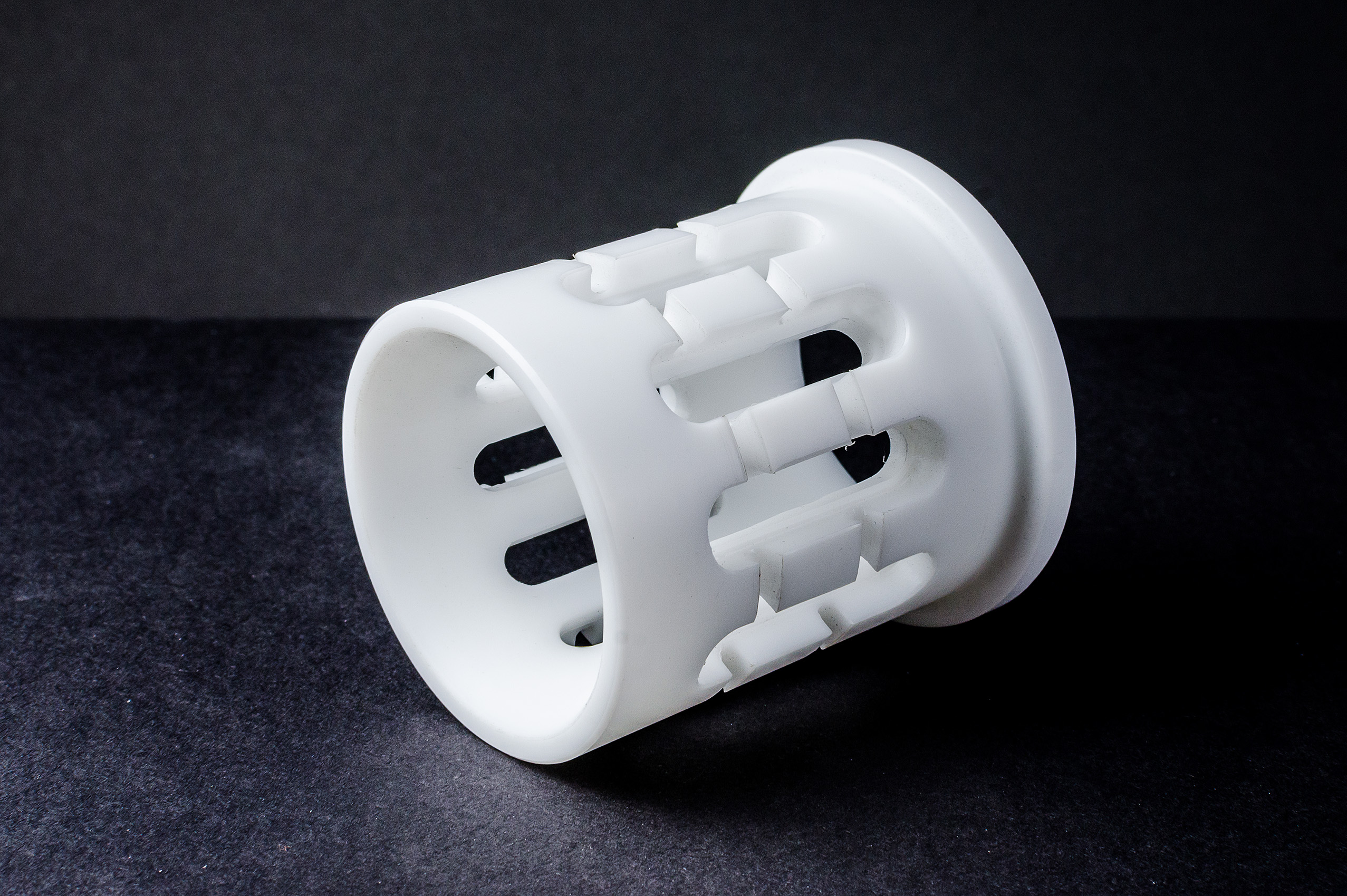A white cylindrical plastic sleeve with elongated slot cut-outs on a dark surface.