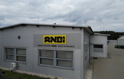 Facade of a grey production hall with the yellow ANBI company logo, windows with blinds and a paved yard.