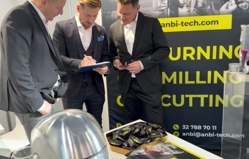 Three men in suits at a trade fair stand talking over a table with a metal part and leaflets.
