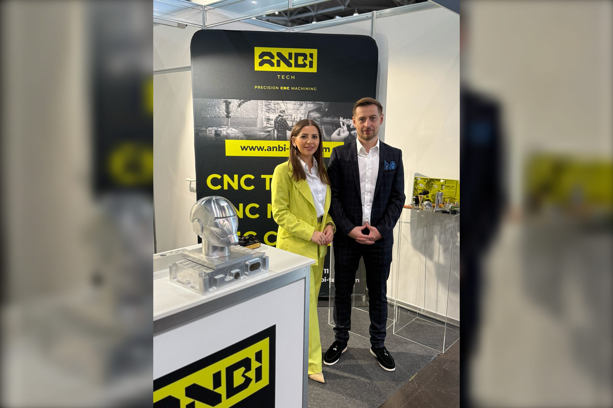 Two people at a trade fair stand next to a displayed metal helmet made by CNC machining and a wall with a logo.