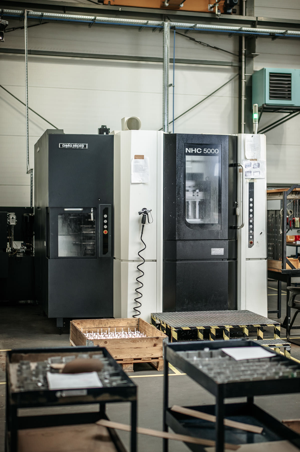 A large black and white CNC machining centre in an industrial hall, with a wooden crate and metal tables in front.
