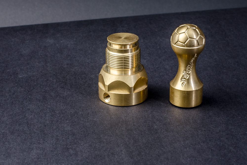 Two precisely turned brass metal parts, one with a thread and the other shaped like a handle with a ball, against a dark background.