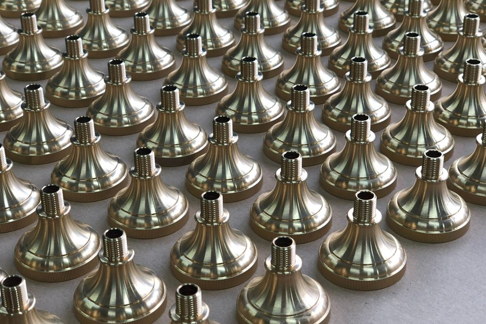 Turned brass parts with a conical shape and a threaded neck, arranged in rows on a grey surface.