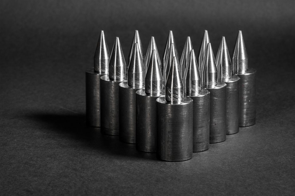 Precisely turned metal components with conical tips, arranged in a group against a dark, uniform background.