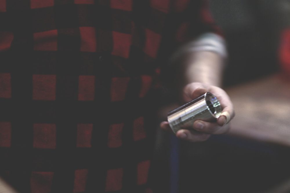 A turned cylindrical metal part held in the hand of a person wearing a red checked shirt against a dark background.