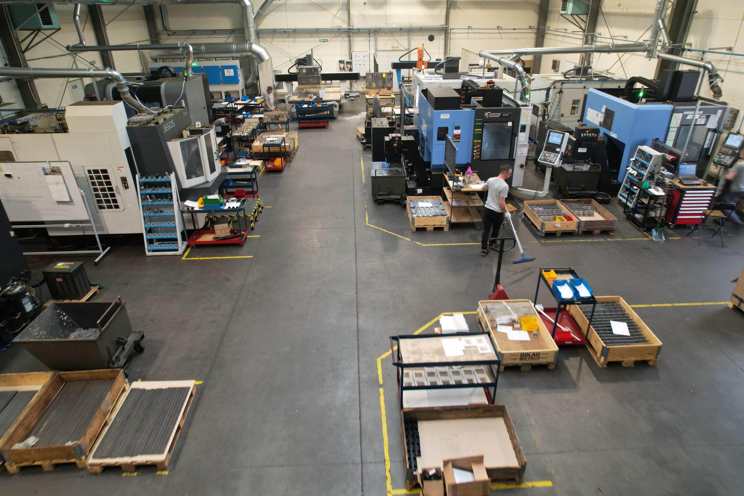 A production hall with numerous CNC machines, pallets full of parts and a worker sweeping the floor.