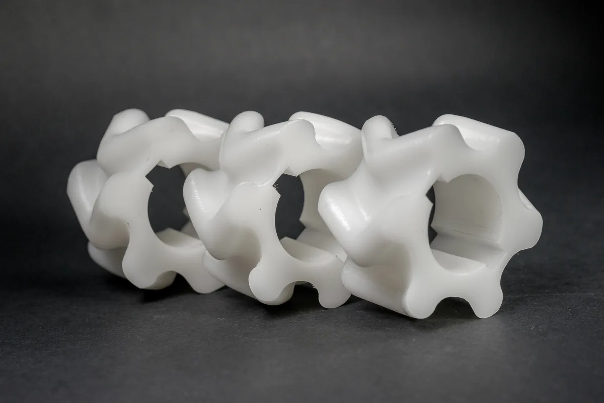 Three white components shaped like twisted gears with holes in the centre, lying in a row against a dark grey background.