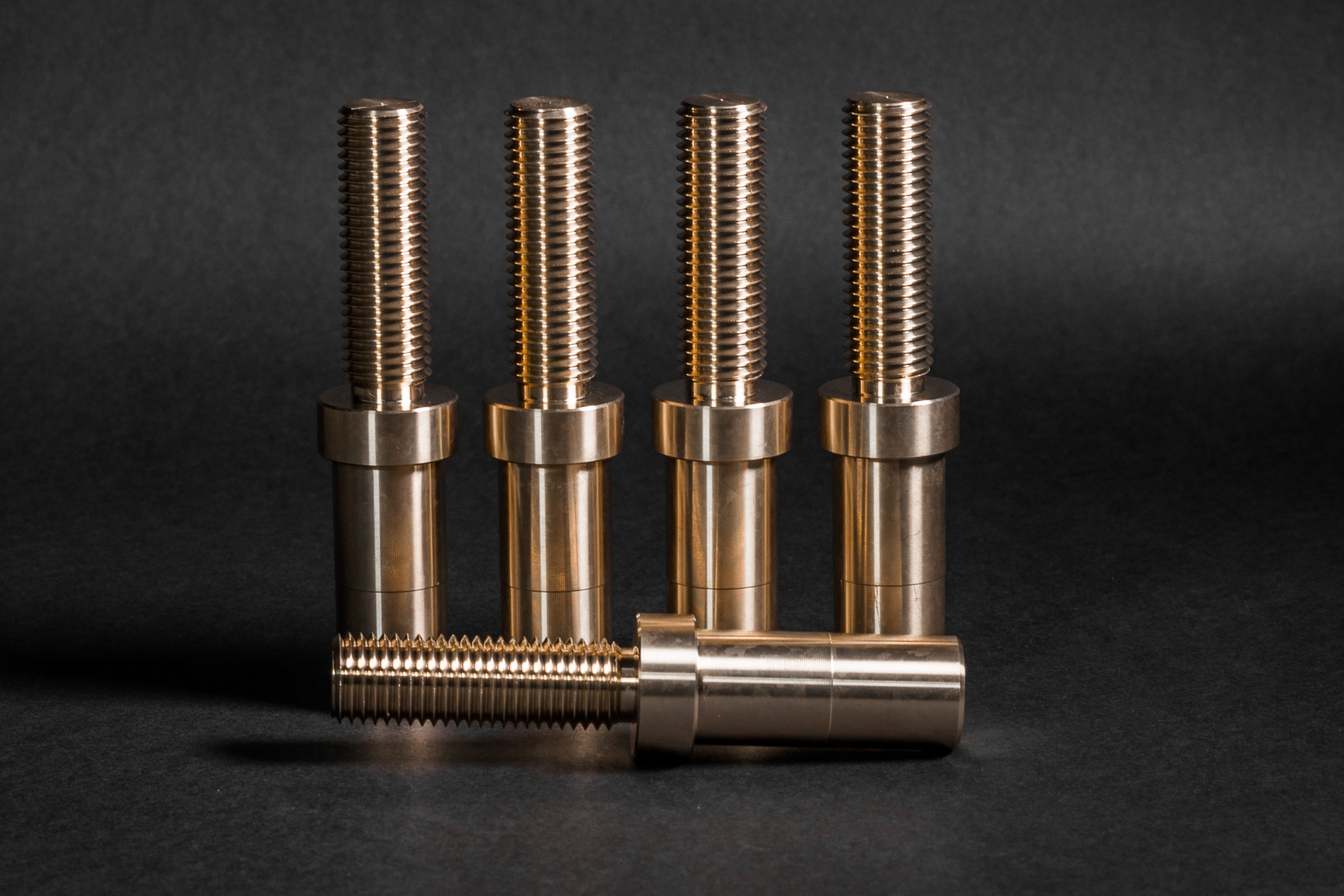 Four brass pins standing upright against a black background, with a fifth identical component lying horizontally in front.
