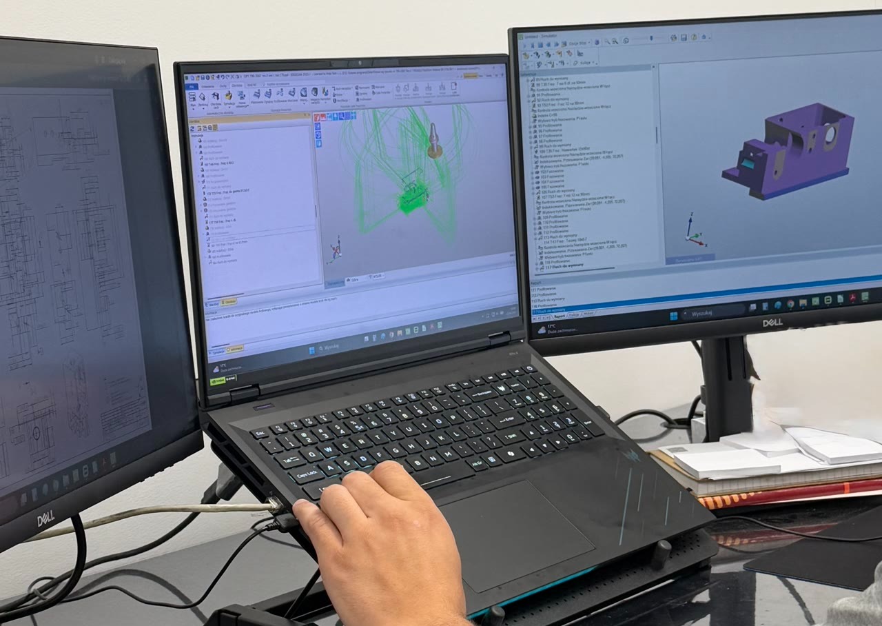 A computer workstation with a laptop and two monitors displaying 3D models and CAD designs for CNC machining.
