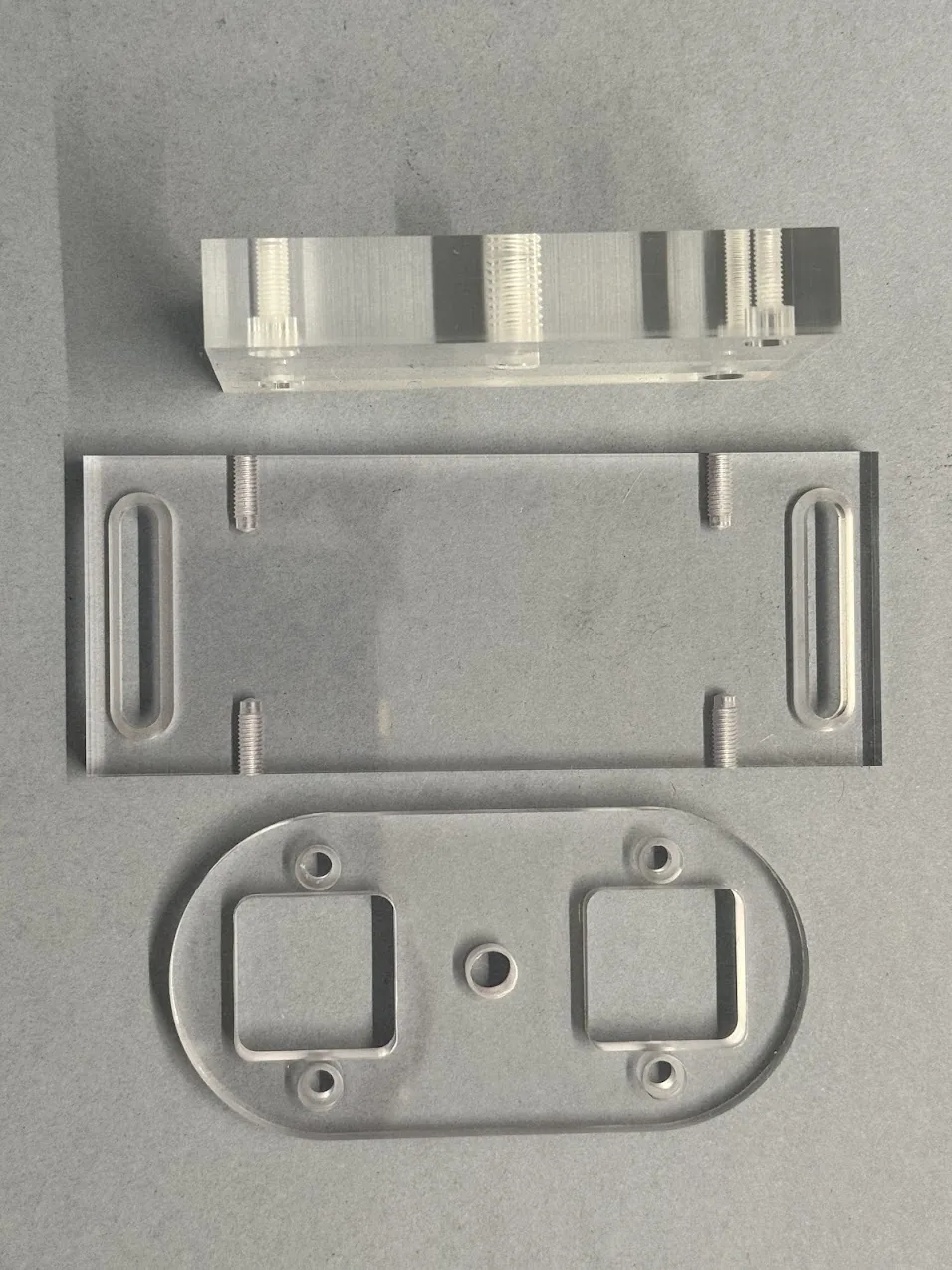 Three colourless, transparent parts with rectangular and oval shapes and cut-out holes against a grey background.