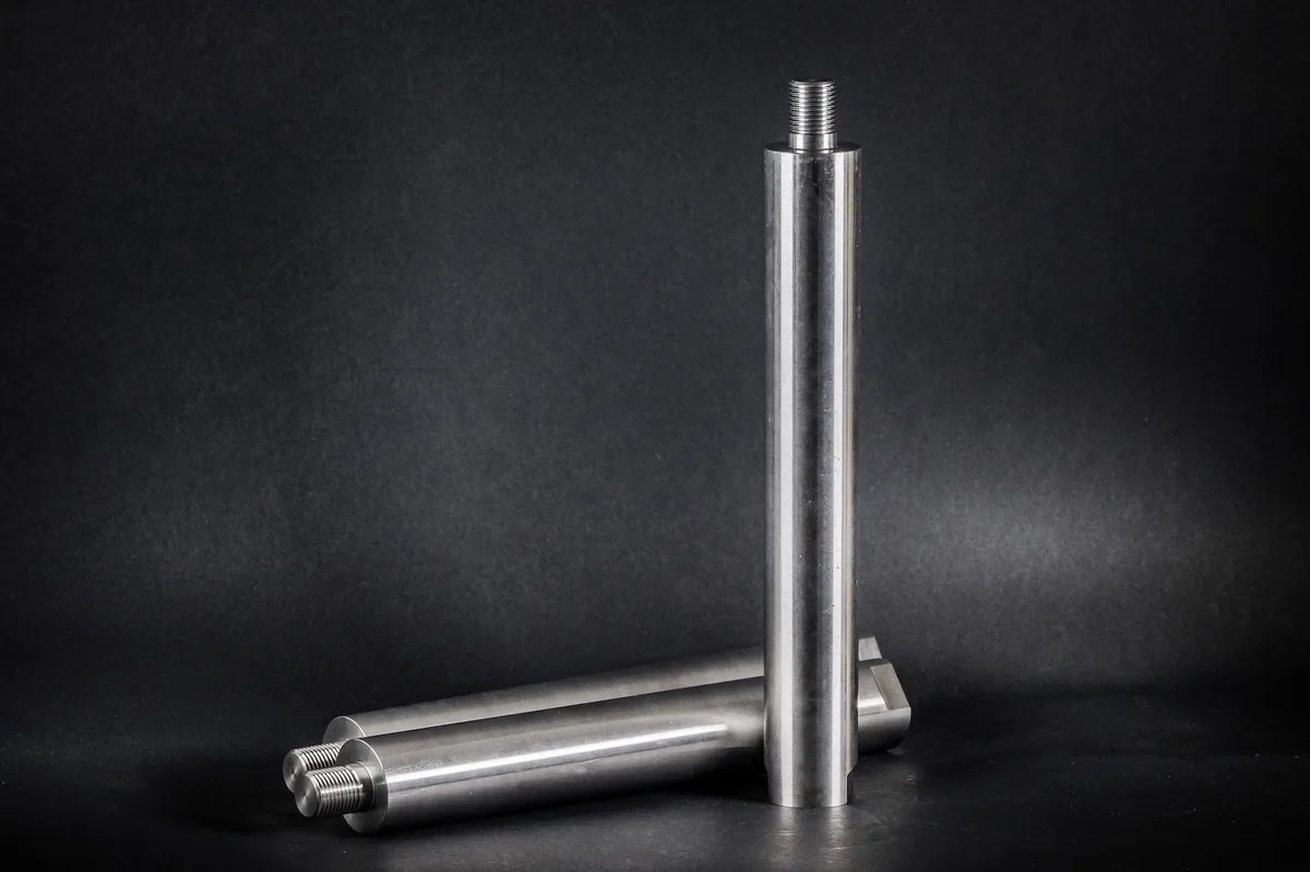 Three turned cylindrical metal shafts with threaded ends, one of them standing upright against a black background.