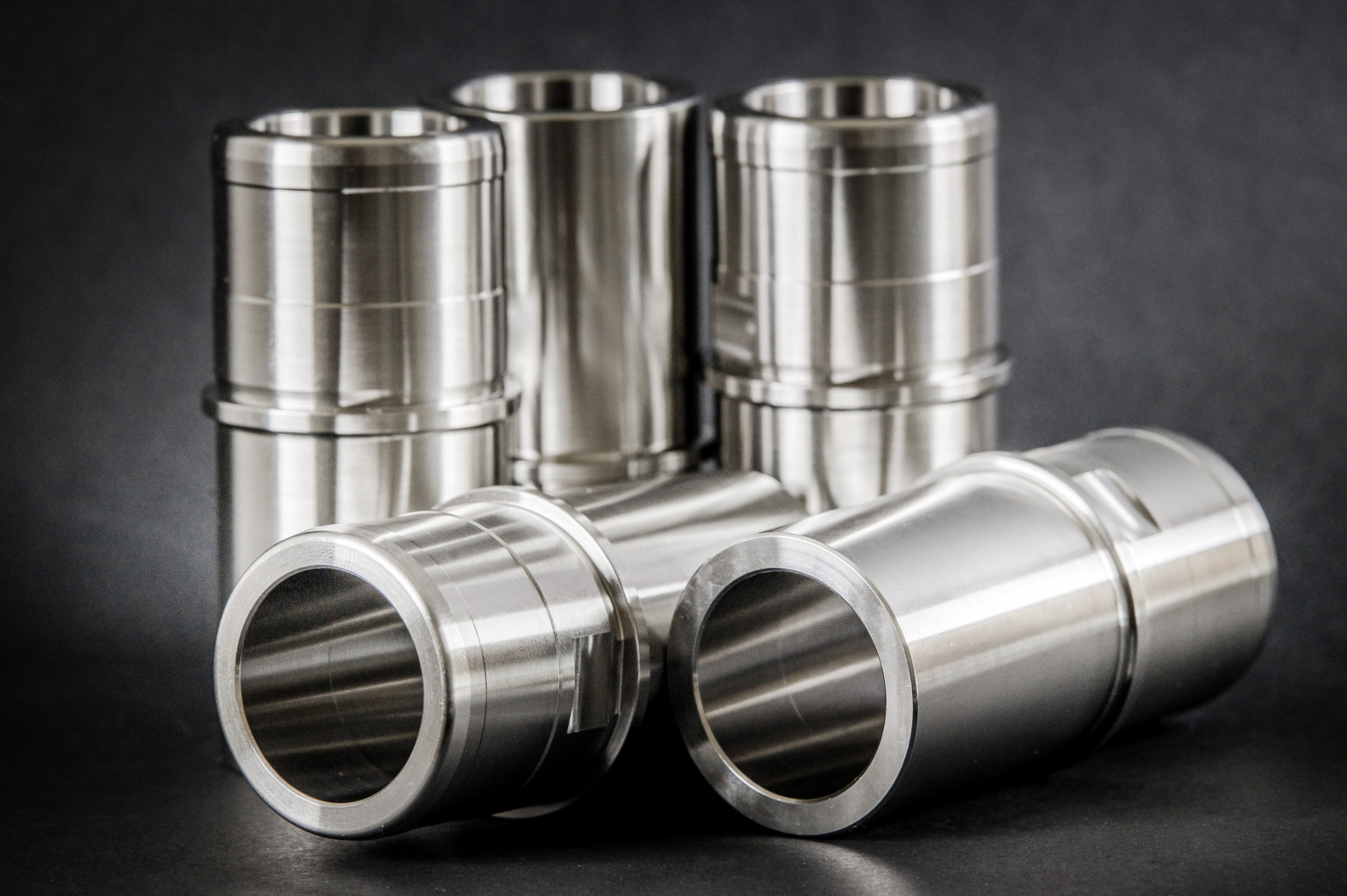 Five precision-turned cylindrical metal bushes with grooves, three upright and two lying on a dark background.