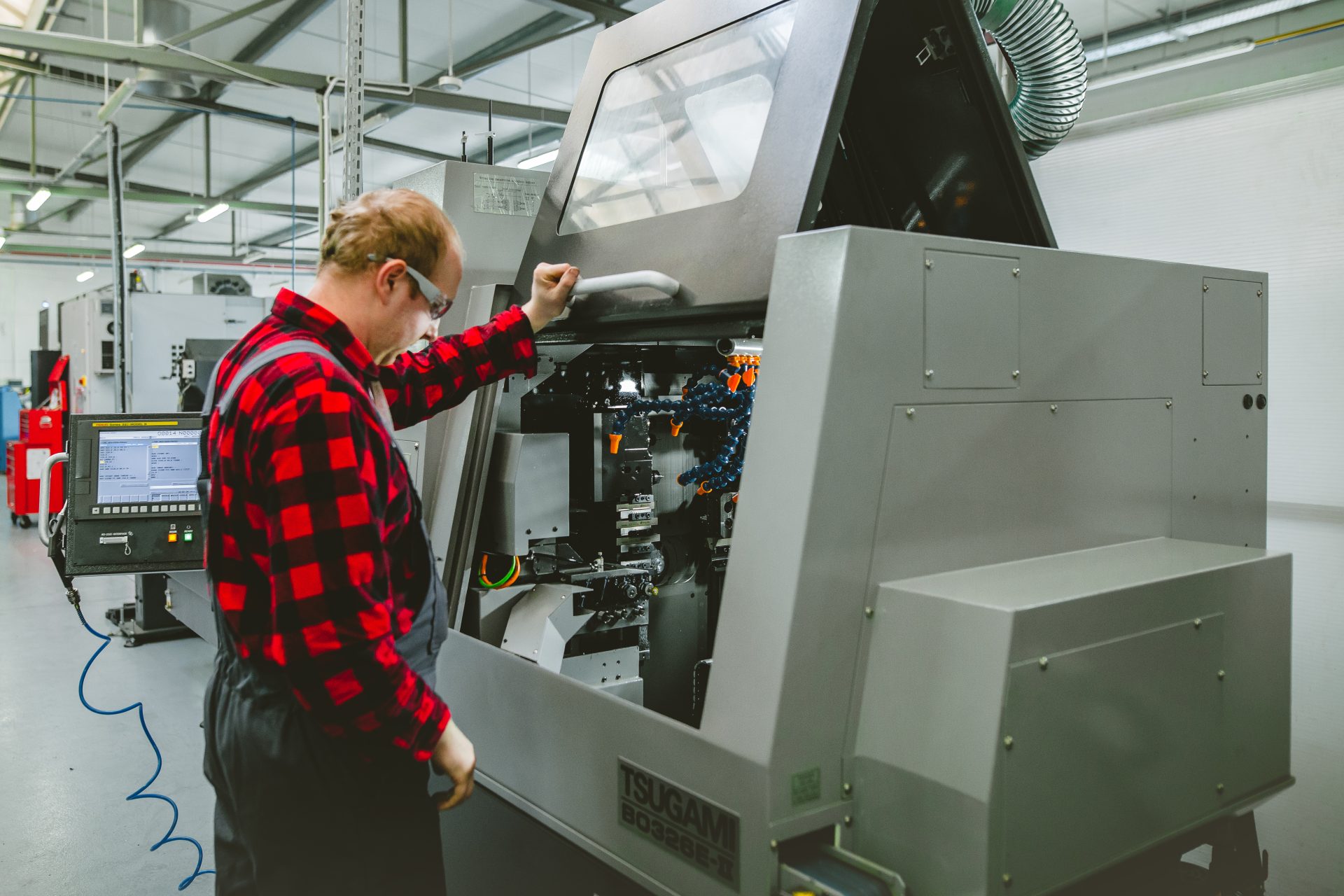 A worker in a red checked shirt operates the panel of an automatic lathe in a bright production hall.
