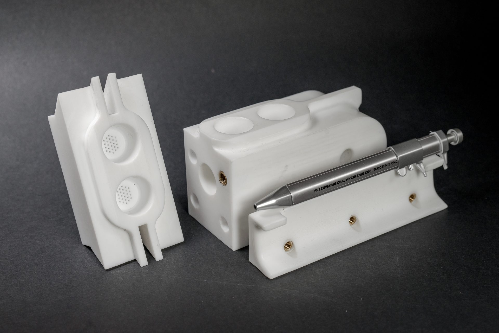 White milled plastic parts with holes and recesses, arranged beside a metal pen to show scale.