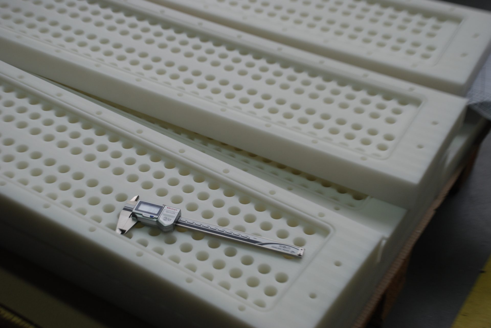 White plastic plates with a dense grid of holes and callipers resting on the surface of a manufactured component.