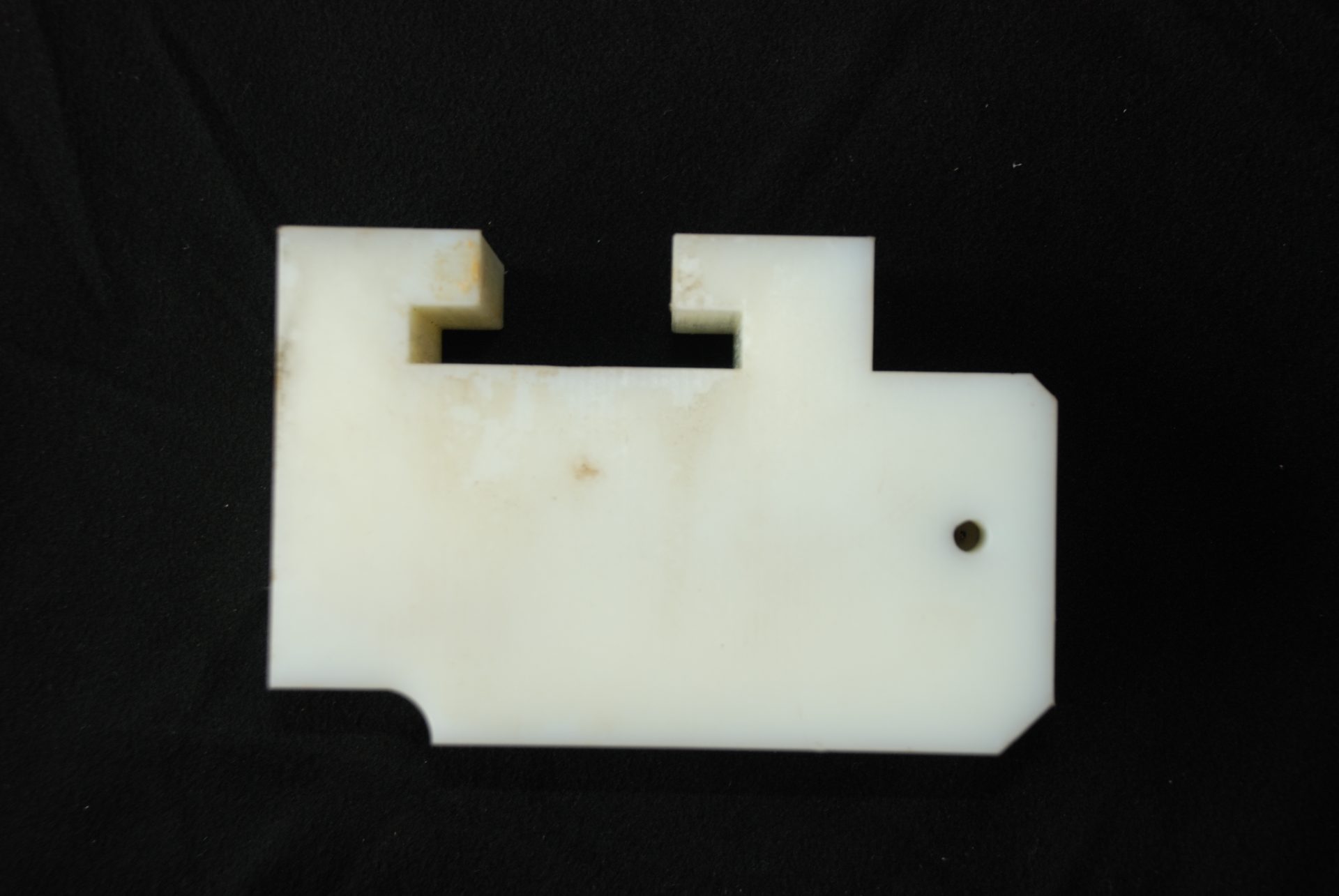 A white milled plastic component with a rectangular cut-out and one hole against a dark studio background.