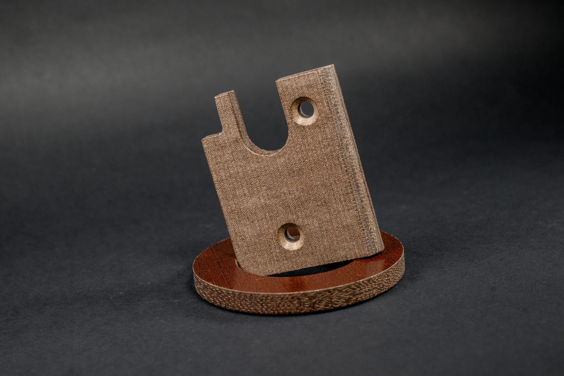 Brown part with two round holes and a semicircular cut-out standing on a round pad with a visible texture.
