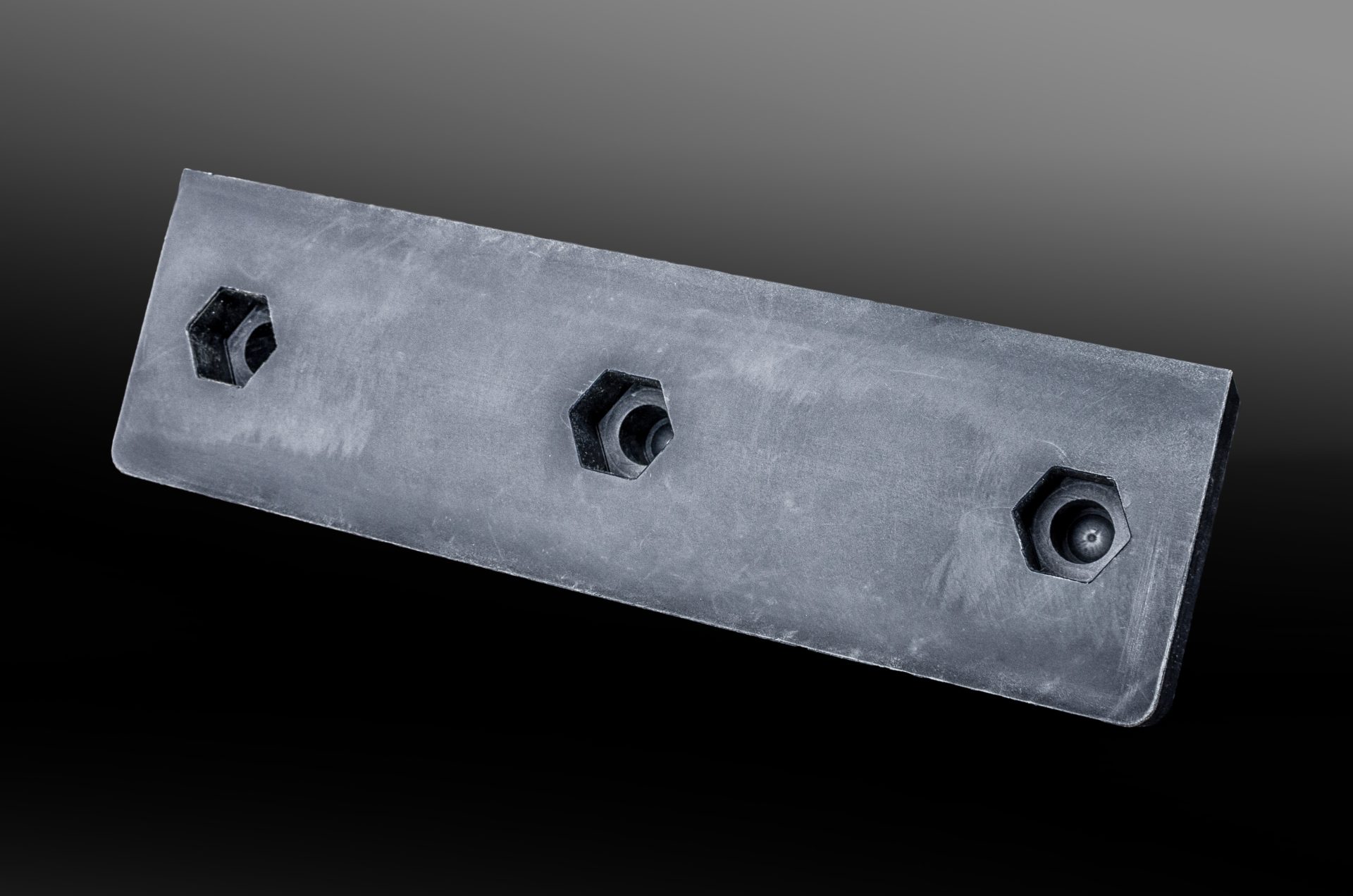 Dark grey elongated plastic plate with three hexagonal mounting holes on a dark background.
