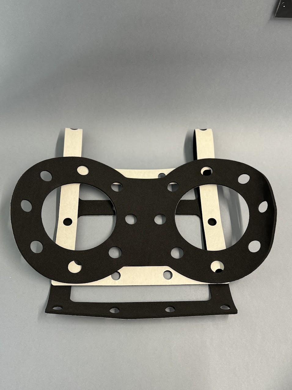 Black double gasket with flange holes, placed on white backing paper on a grey background.