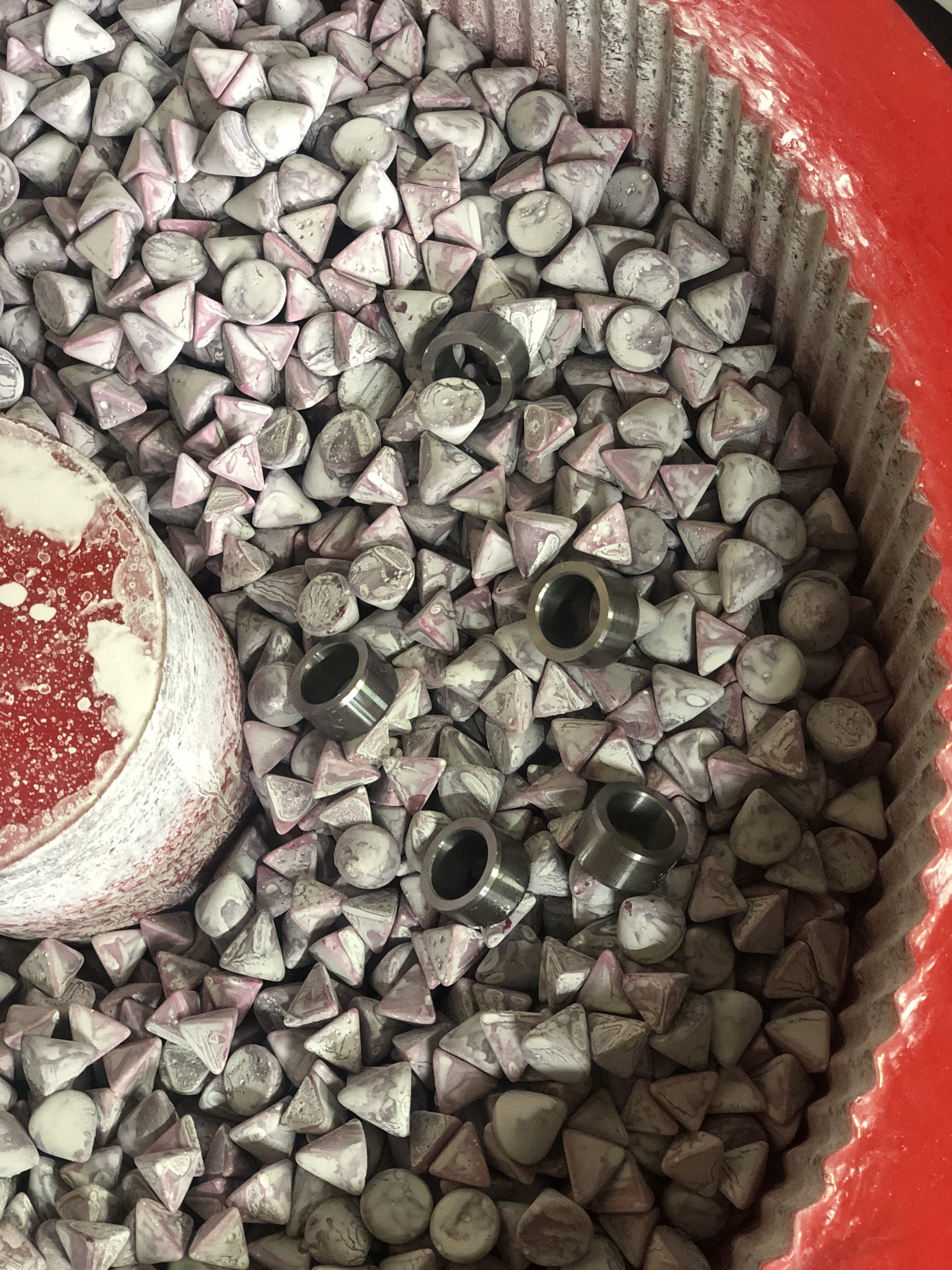 Metal parts in a red bowl with abrasive media during surface smoothing among ceramic shapes.