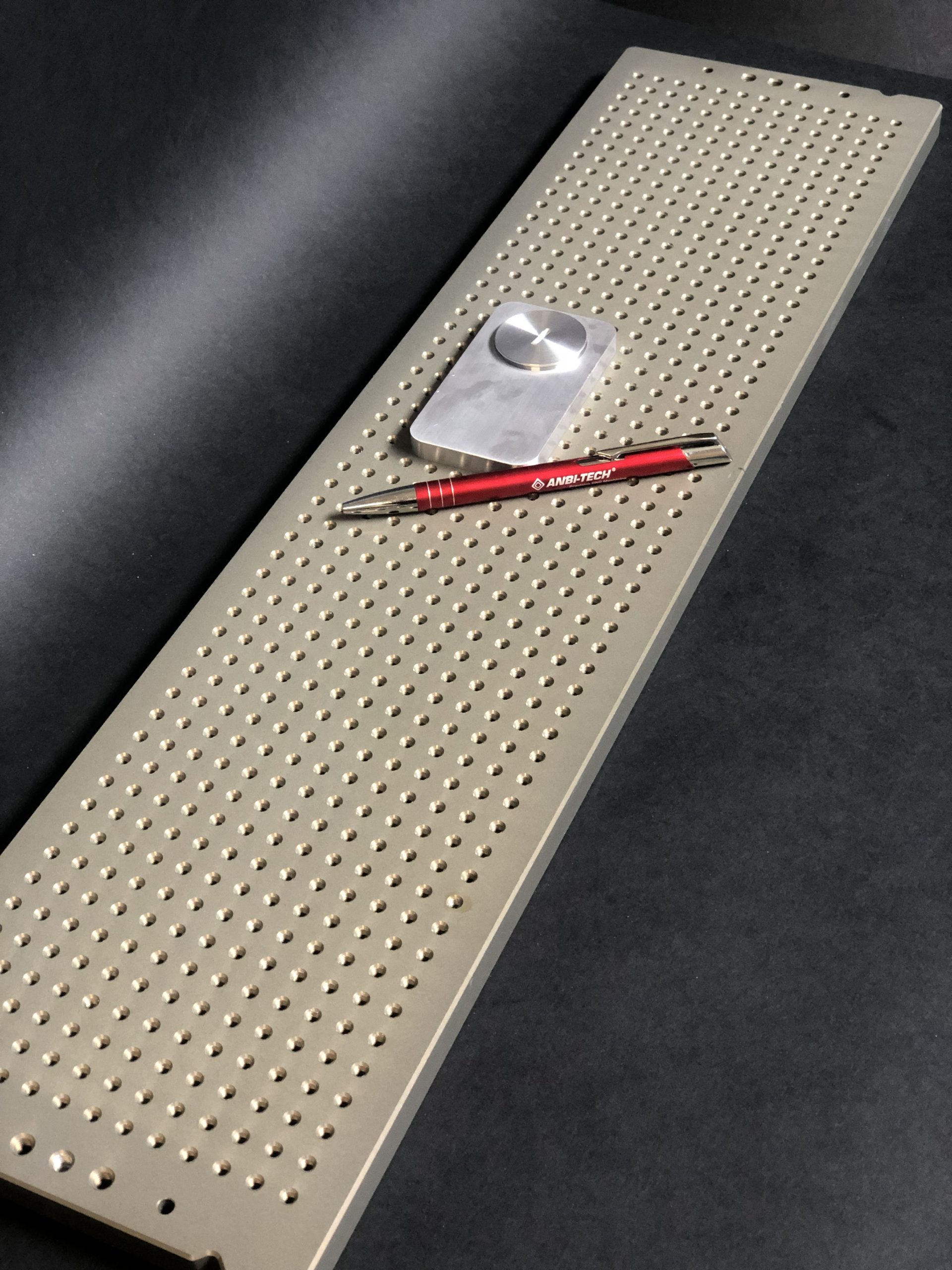 Long metal mounting plate with a regular grid of holes, a small block and a red pen on a dark background.
