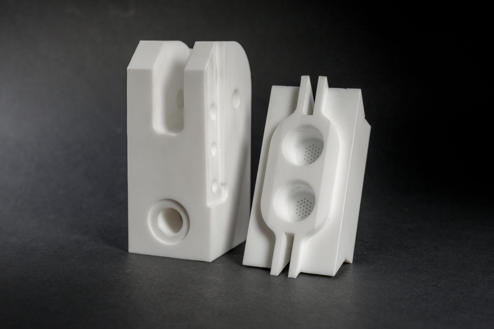 Two white milled plastic parts with holes and deep recesses standing against a dark studio background.