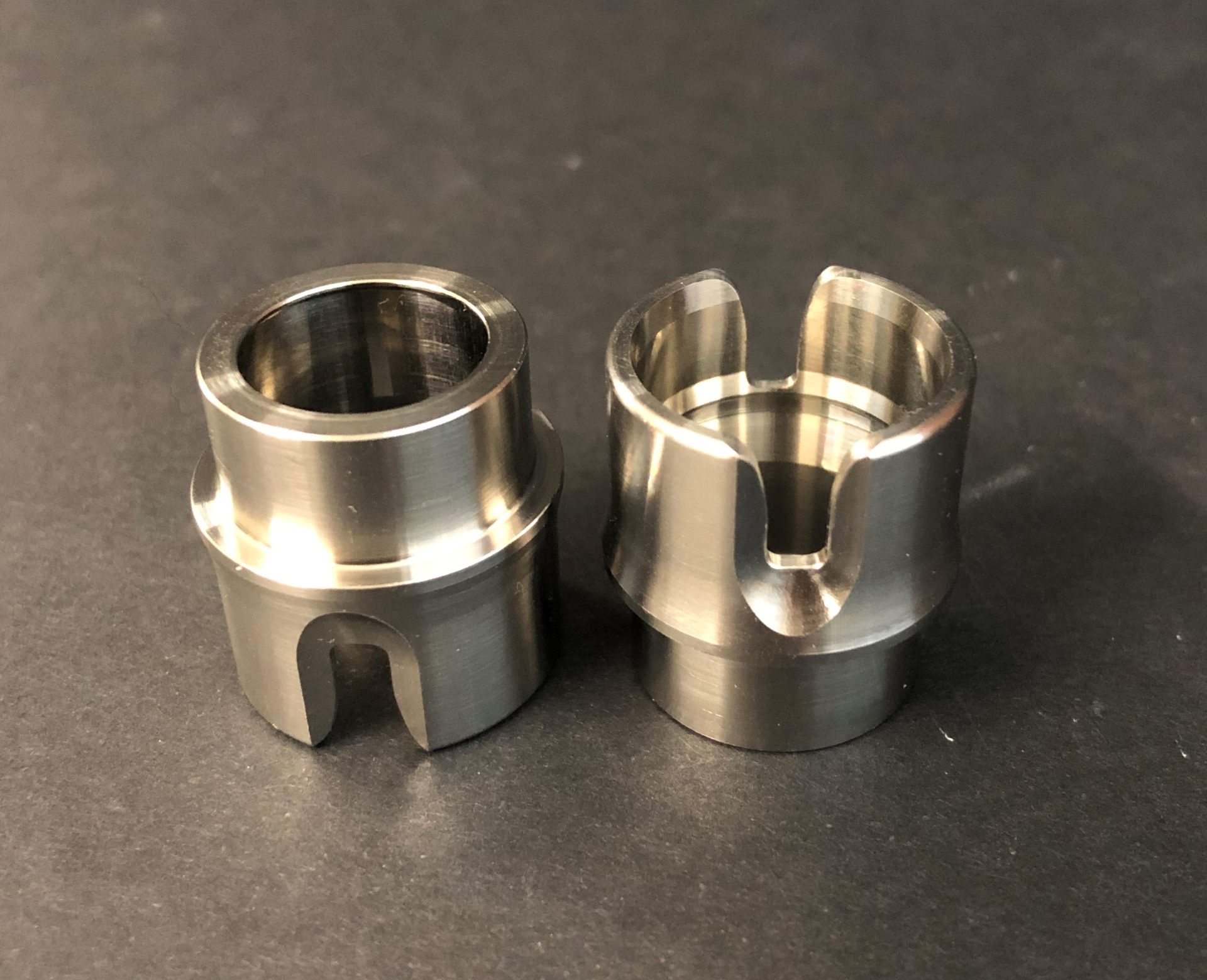 Two small metal parts of cylindrical shape standing against a dark background, one of them with a cut-out at the top.
