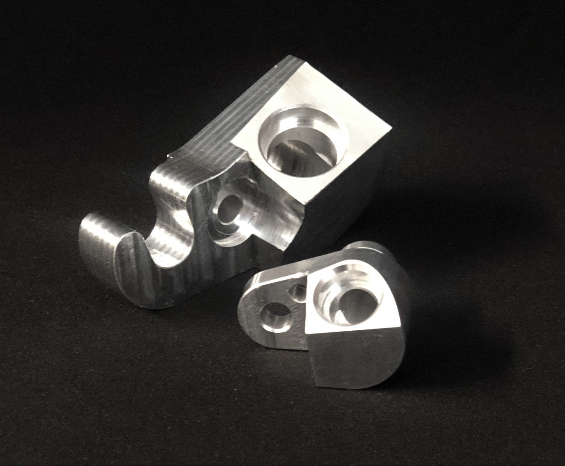 Two silver metal components with milled surfaces and holes lying on a matt black background.