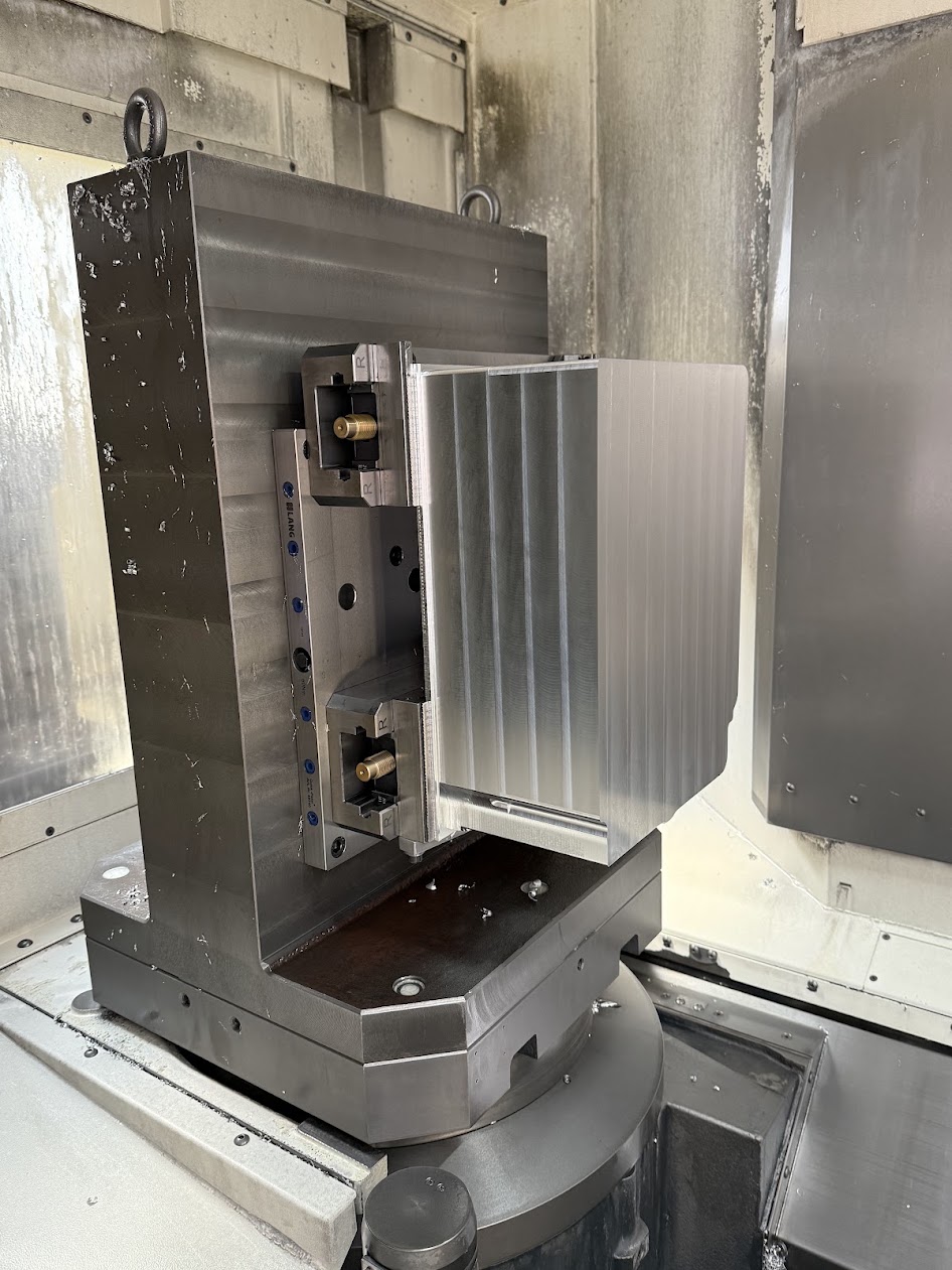 A light-coloured ribbed block is clamped to a vertical plate inside a dark grey metal machine tool.