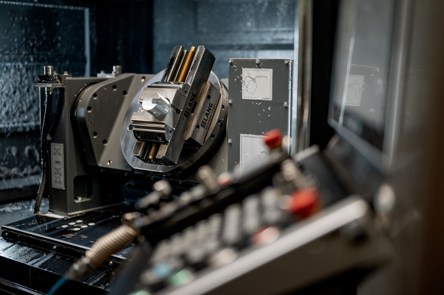 Metal vice with a clamped workpiece inside the machining chamber, seen from behind a control panel with buttons.